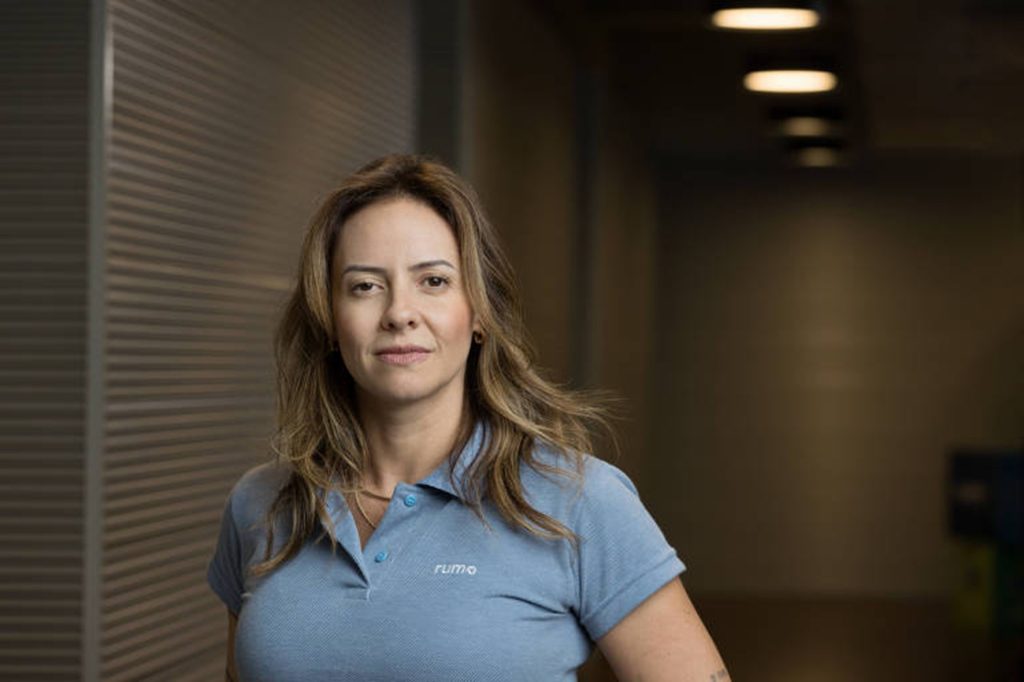 Mulher com cabelo solto, usando uma camisa polo azul com o logo 'rumo'. Ela está em um ambiente interno, com uma expressão séria e um fundo desfocado.