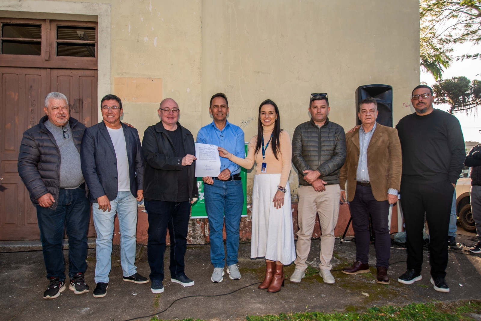 Grupo de pessoas posando para uma foto em frente a um prédio antigo. Eles estão segurando um documento.