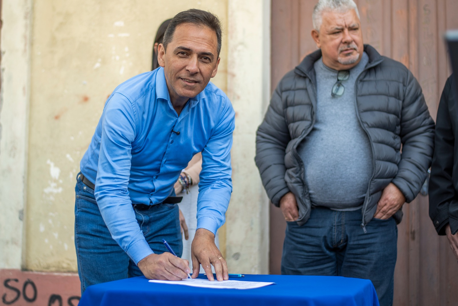 Homem assinando um documento em uma mesa azul, com outro homem ao lado. A cena parece ser parte de uma cerimônia ou evento.