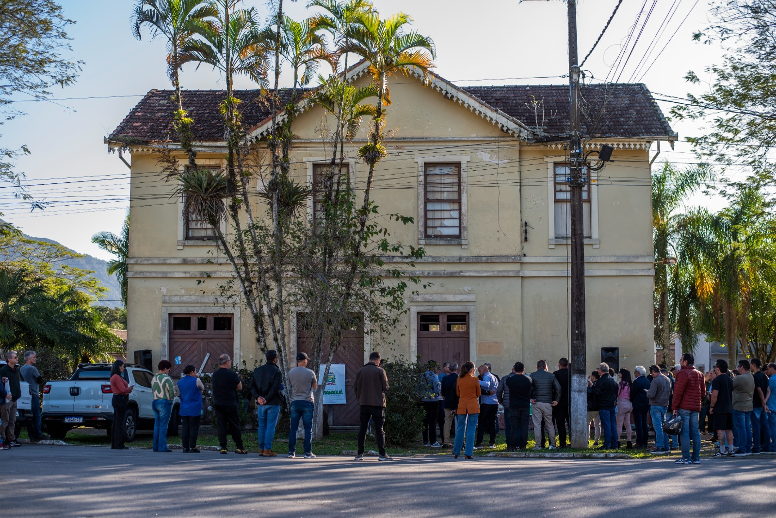 Grupo de pessoas reunidas em frente à estação de trem "Alexandra". Há carros estacionados e vegetação ao redor.