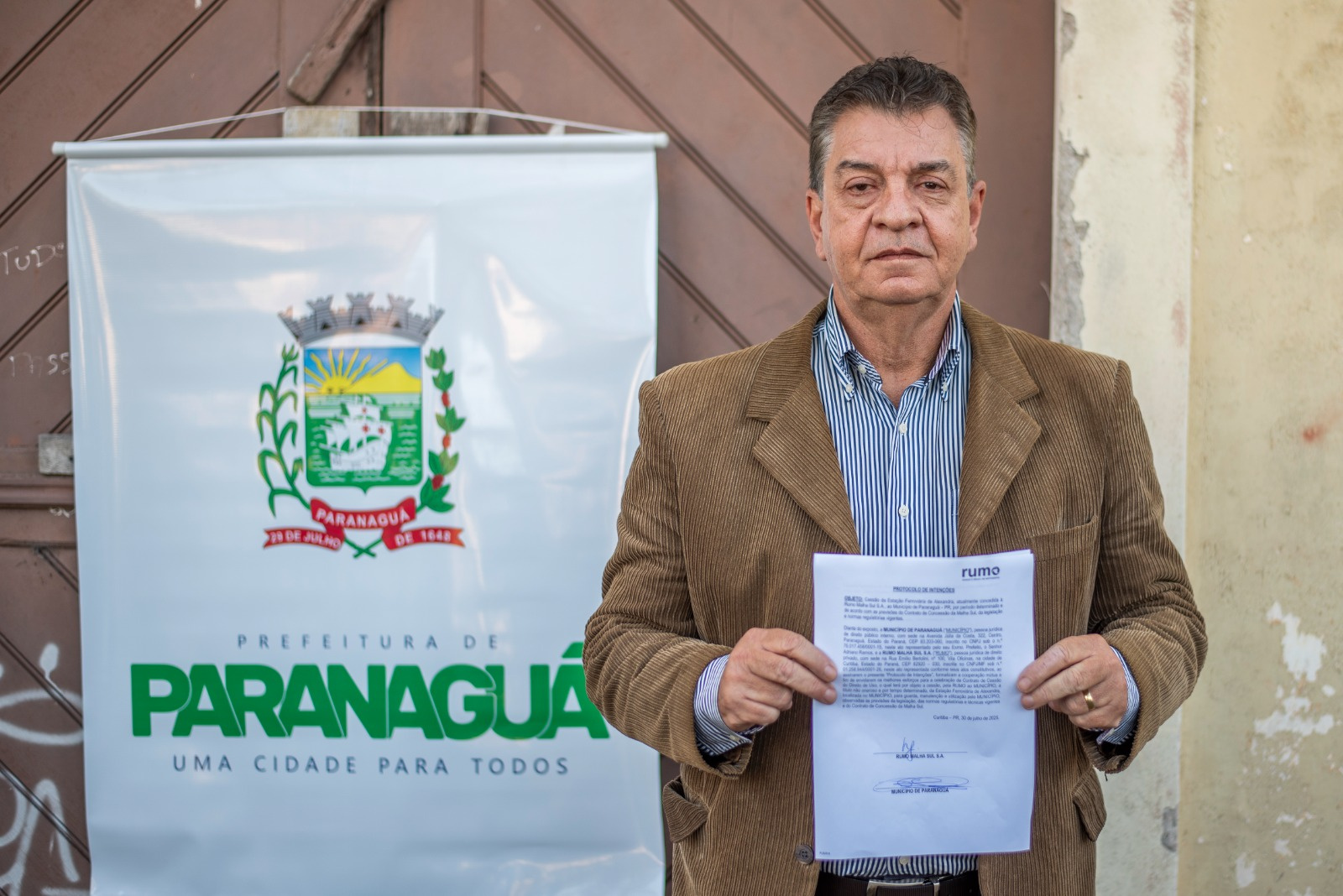 Homem segurando um documento e em pé ao lado de uma placa da Prefeitura de Paranaguá.