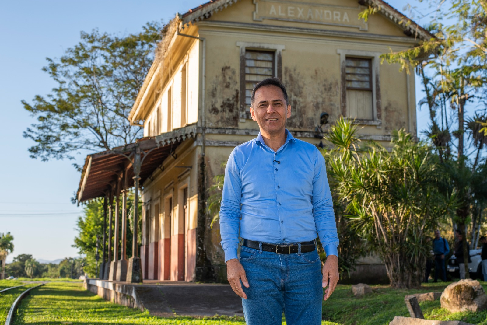 Homem de camisa azul e calça jeans em frente à estação de trem "Alexandra". A estação tem uma aparência antiga e está cercada por vegetação.