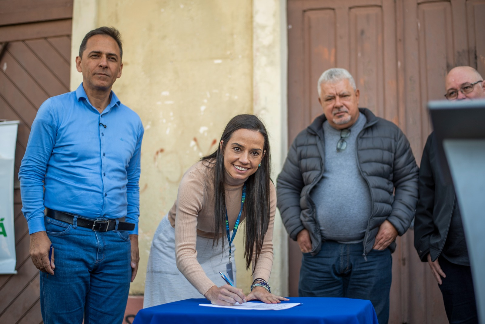 Mulher sorrindo enquanto assina um documento em uma mesa azul, com dois homens ao lado. O fundo é uma parede desgastada.