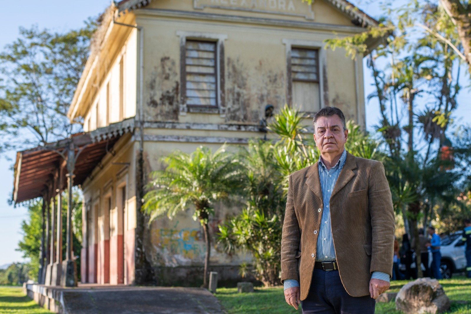 Homem de terno marrom em frente a uma estação de trem antiga com a palavra "Alexandra" acima das janelas. A estação está um pouco desgastada, com vegetação ao redor.