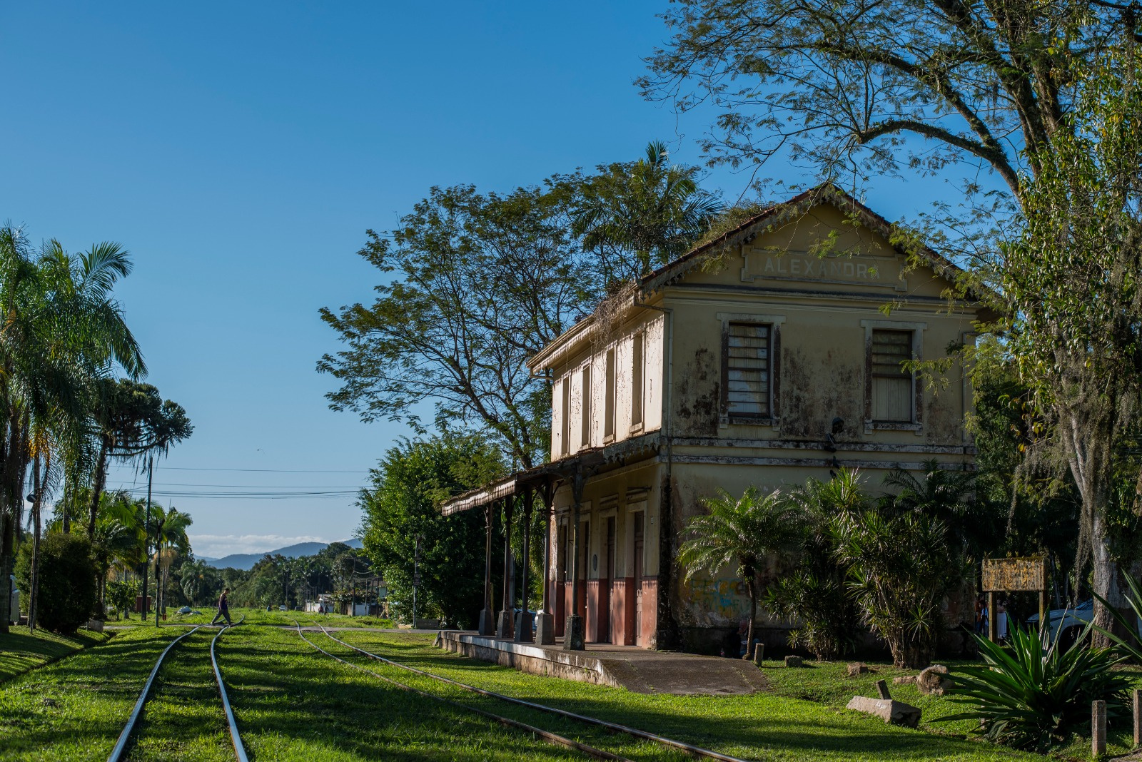 Vista da antiga estação de trem "Alexandra" com trilhos em primeiro plano, vegetação exuberante e céu azul. A estação mostra sinais de desgaste.