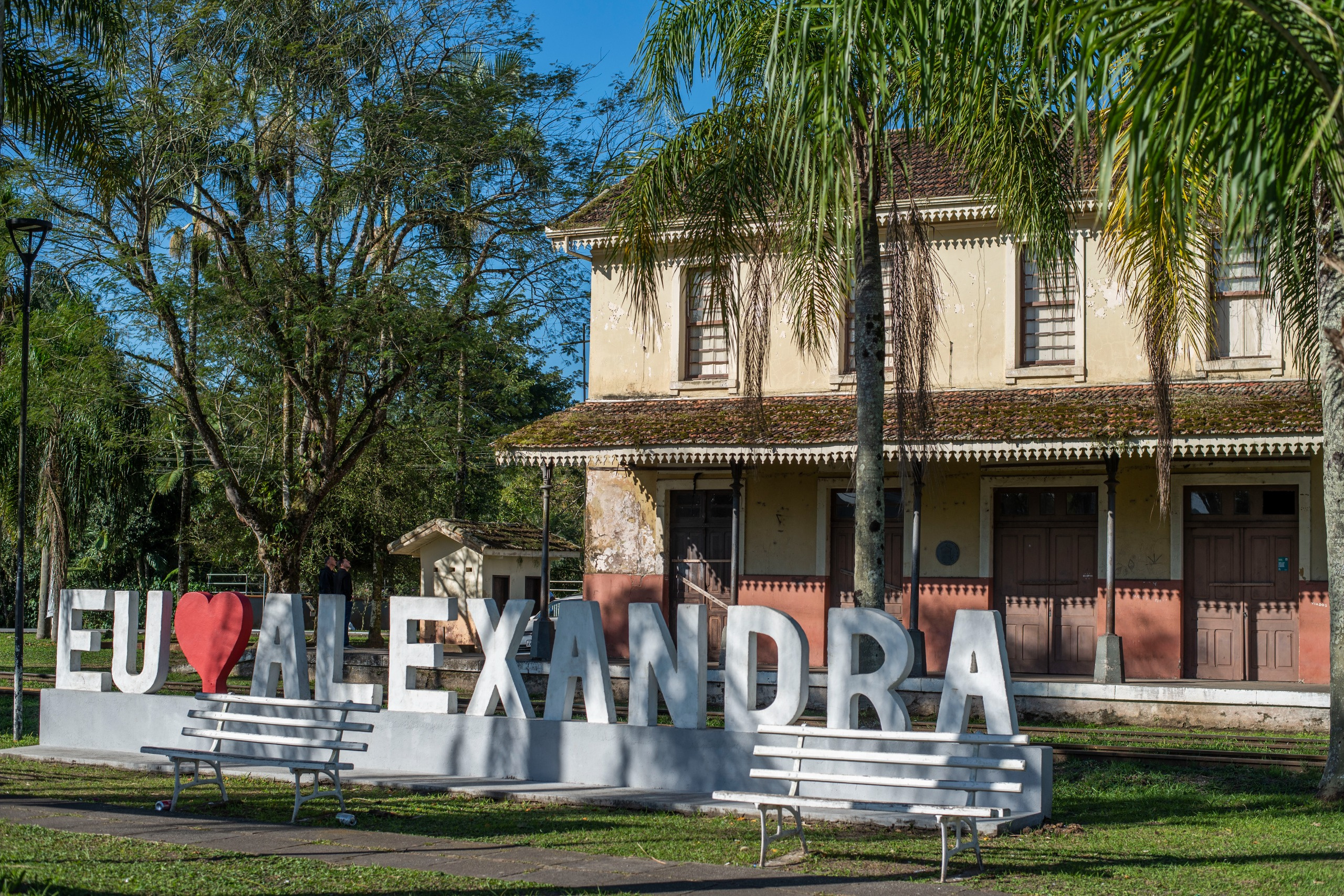 Paisagem da antiga estação de trem "Alexandra", com um letreiro decorativo em primeiro plano que diz "EU AMO ALEXANDRA". O letreiro é composto por letras brancas grandes e um coração vermelho, e há dois bancos brancos em frente a ele. A estação está ao fundo, cercada por árvores e vegetação.