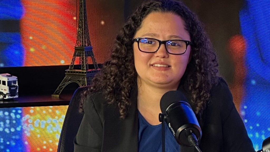 Pessoa falando ao microfone em um estúdio com objetos decorativos ao fundo.