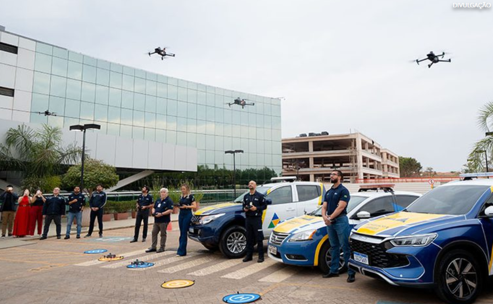 Equipe de segurança junto a carros oficiais enquanto vários drones voam sobre um pátio em frente a um prédio de vidro.