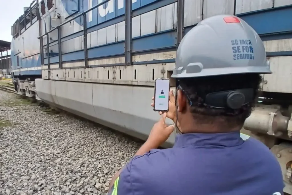 Trabalhador de costas com capacete segurando um celular enquanto inspeciona um equipamento ferroviário sobre trilhos.