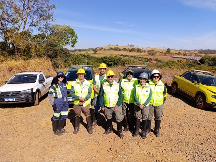 Um grupo de sete pessoas, a maioria vestindo coletes de segurança amarelos e capacetes, está em pé em uma estrada de terra sob um céu azul, com dois veículos e uma paisagem campestre ao fundo.