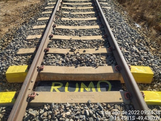 Vista de cima dos trilhos de trem, com dormentes de concreto e brita. Um dormente central tem a palavra "RUMO" em amarelo.