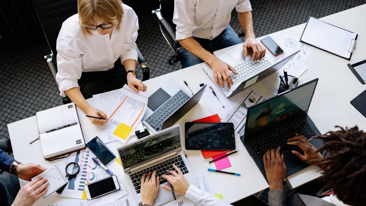 Pessoas trabalhando ao redor de uma mesa com laptops, papéis e celulares.
