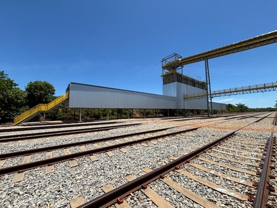 Área ferroviária com trilhos múltiplos ao lado de estrutura industrial metálica sob céu azul.