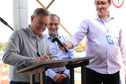 Três homens durante um evento, com um deles assinando um documento enquanto outro segura um microfone.