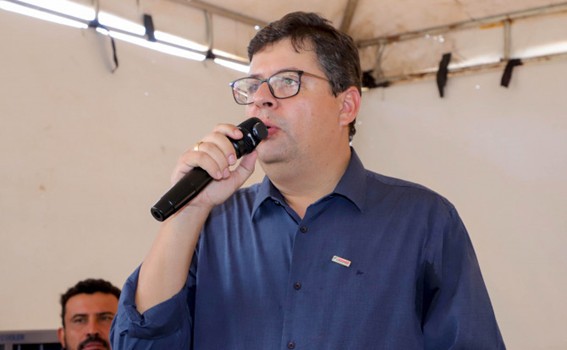 Homem de óculos falando ao microfone durante um evento coberto, vestindo camisa social azul. Ao fundo, outra pessoa observa.