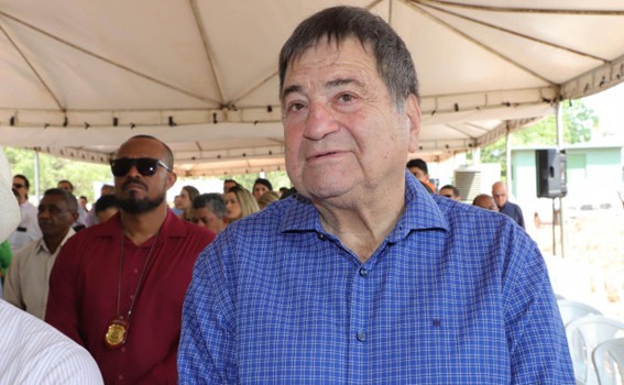 Homem de camisa xadrez azul em primeiro plano, de pé sob tenda aberta, com público sentado e em pé ao fundo num ambiente iluminado