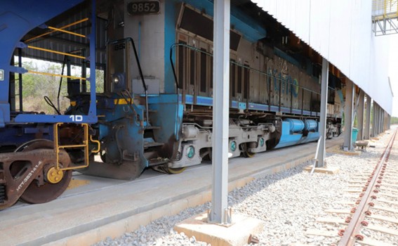 Vista lateral de trem azul estacionado sobre trilhos, parcialmente abrigado por cobertura metálica; piso de concreto e lastro de pedras ao redor.