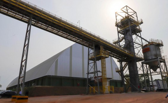 Galpão de telhado triangular revestido em chapas metálicas, com correia transportadora elevada e torre metálica de silos ao lado, sob forte luz do sol.