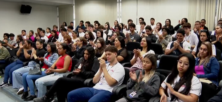 Plateia com dezenas de jovens e adultos sentados em um auditório, assistindo a uma apresentação e aplaudindo, com cadeiras organizadas em fileiras.