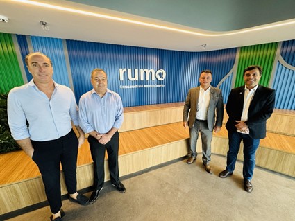 Quatro homens sorridentes em pé, posando para uma foto em um escritório moderno. Atrás deles, há uma parede ondulada azul com o logotipo da empresa 'Rumo'