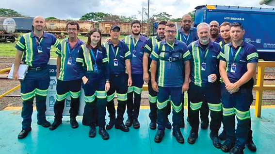 Grupo de cerca de doze colaboradores posando para foto em área externa de pátio ferroviário. Todos usam uniforme azul-marinho com faixas refletivas amarelas e crachás. Ao fundo, vagões de trem e vegetação.