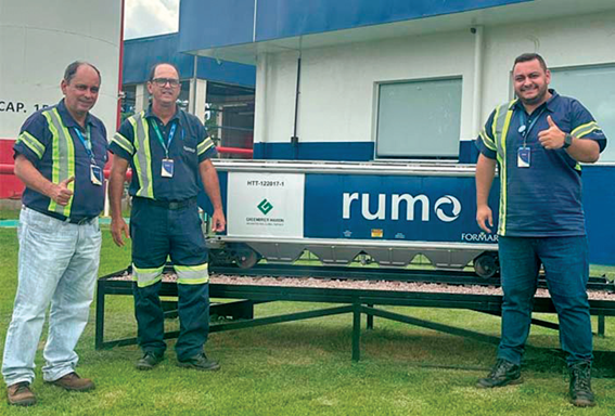 Três homens com uniformes azuis e coletes refletivos posam sorrindo em frente a um vagão ferroviário com a marca Rumo. Um deles faz sinal de positivo com o polegar. O ambiente é externo, com grama e estrutura da empresa ao fundo.