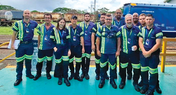 Grupo de cerca de doze colaboradores posando para foto em área externa de pátio ferroviário. Todos usam uniforme azul-marinho com faixas refletivas amarelas e crachás. Ao fundo, vagões de trem e vegetação.