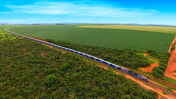 Vista aérea de um longo trem azul atravessando uma área rural, entre vegetação nativa e extensas plantações verdes, sob céu azul claro. O trilho acompanha uma estrada de terra vermelha e se curva ao longe na paisagem.