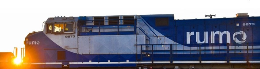 Locomotiva azul da Rumo vista de lado, com o sol baixo ao fundo iluminando a cena.