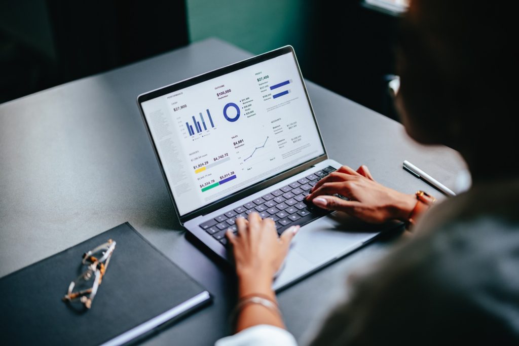 Pessoa usando um notebook com gráficos e indicadores financeiros na tela, sobre uma mesa de trabalho.