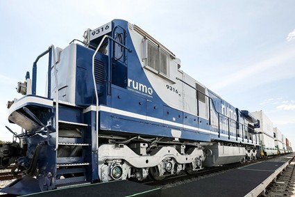 Locomotiva azul e branca da Rumo, vista em perspectiva de baixo para cima, sobre trilhos em um ambiente aberto.