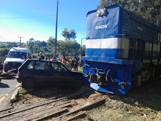 Locomotiva azul de número 4404 colidida com um carro Fiat Uno escuro sobre trilhos de trem, com pessoas observando ao fundo.